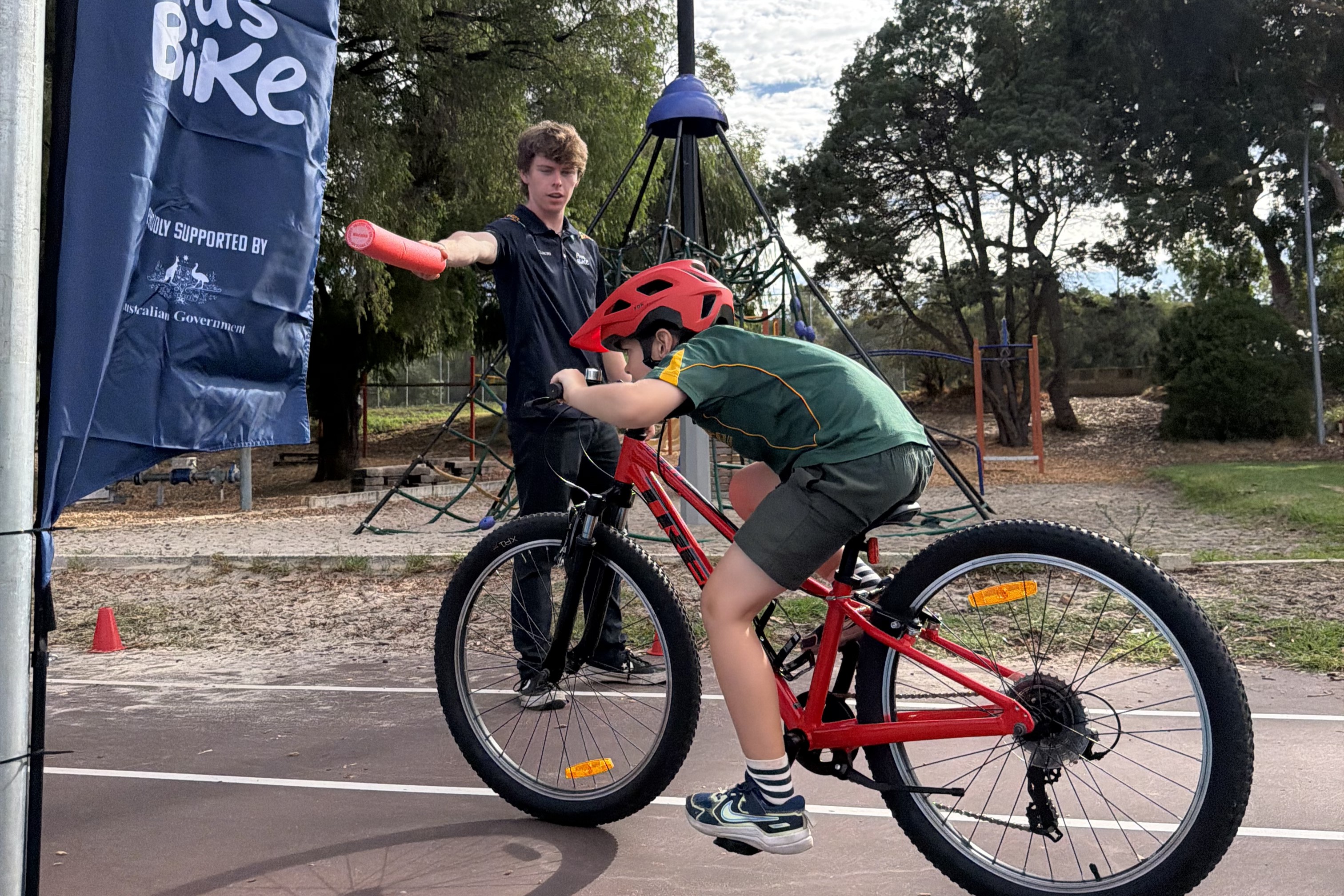 A student on a bicycle rides under a pool noodle held by an AusBike instructor as part of Ride2School Day activities at Kensington Primary School in Western Australia
